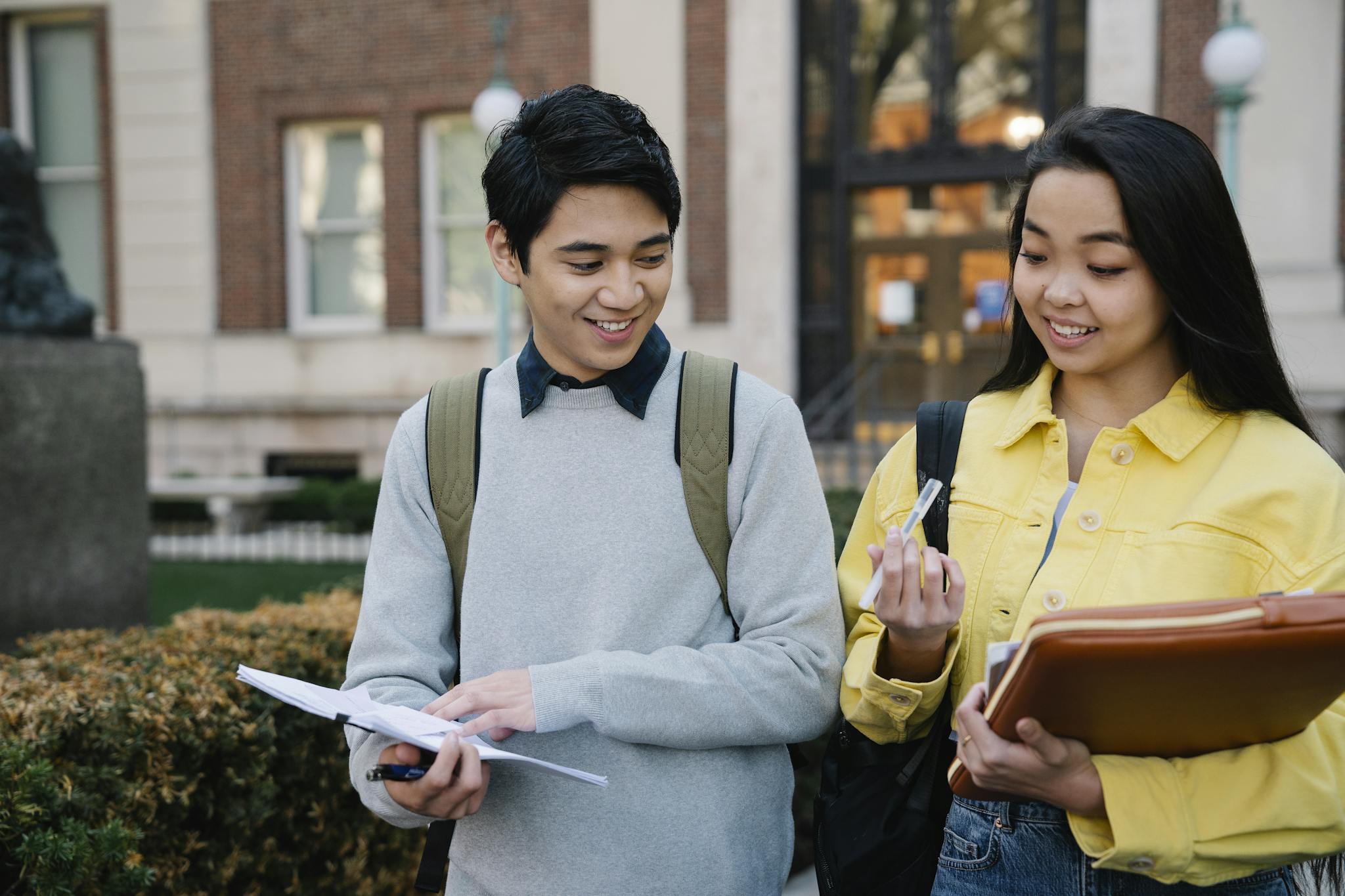 Two students collaborate outdoors on a college campus, sharing smiles and study materials.
